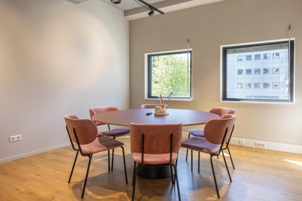 Modern meeting room on Overschiestraat with a round table, six pink chairs, and large windows.
