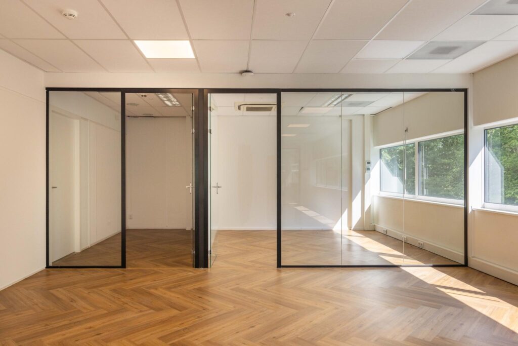 Modern office space with glass partition walls, wooden herringbone floor, and large windows letting in natural light.