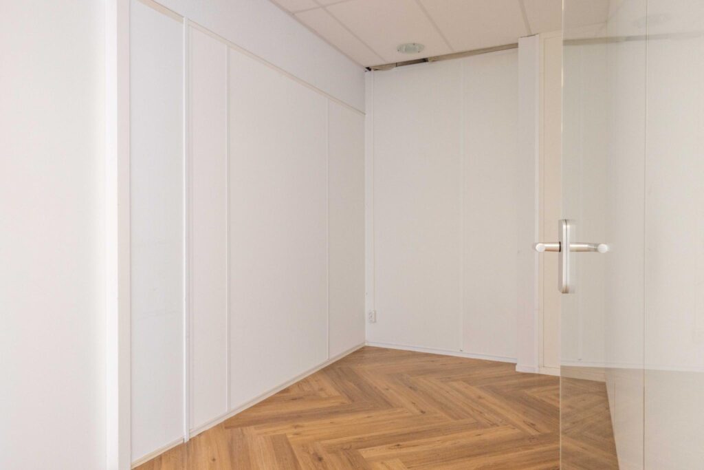 Empty white office room with herringbone wooden flooring and a glass door.