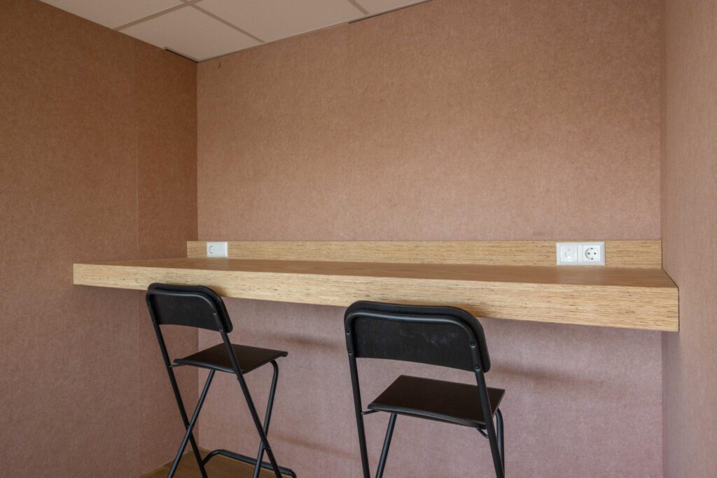Minimalist workspace with a wooden bar-height desk, two black chairs, and wall-mounted power outlets.