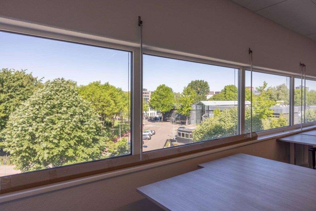 View from an office window overlooking Overschiestraat with trees, parked cars, and industrial buildings.