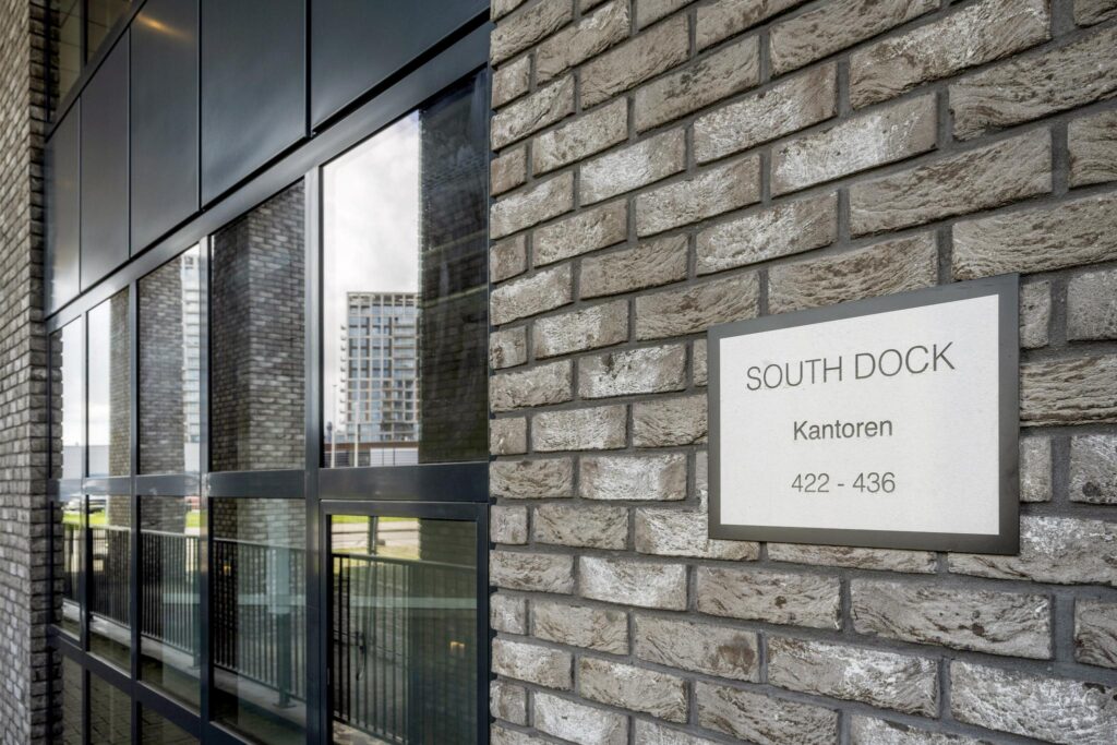 Sign reading “South Dock Kantoren 422 - 436” mounted on a brick wall next to office building windows.