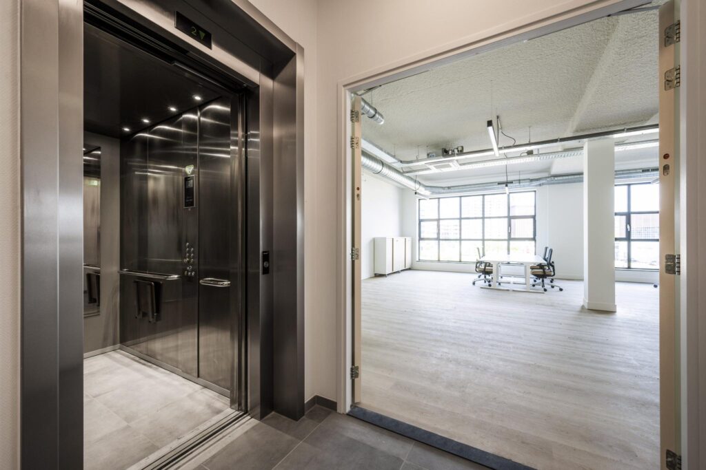 Modern elevator and entrance to a bright, industrial-style office space with large windows and minimal furnishings.