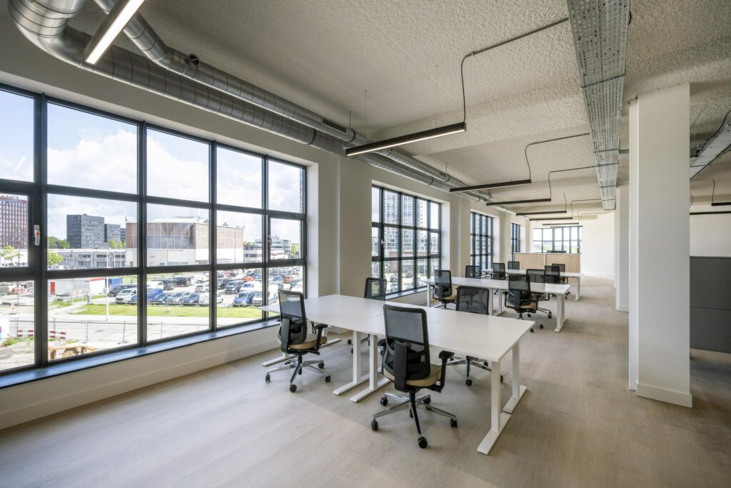 Modern open-plan office space with large windows and multiple white desks and office chairs.