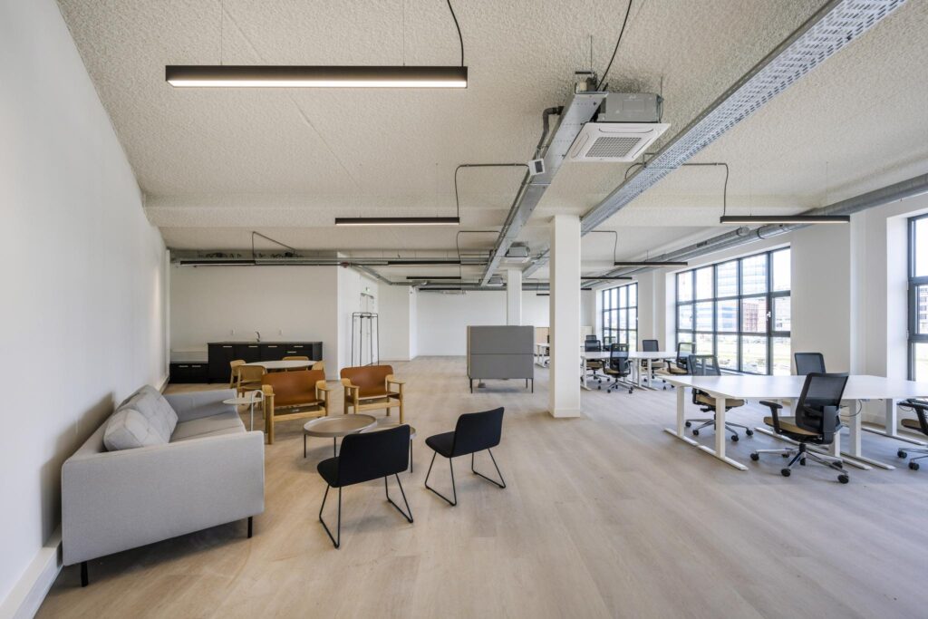 Modern open-plan office space with shared desks, lounge area, and large windows providing natural light.