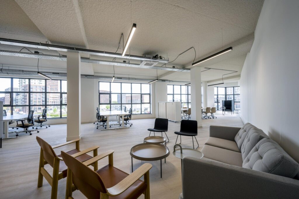 Modern open-plan office space with large windows, workstations, and a casual seating area.