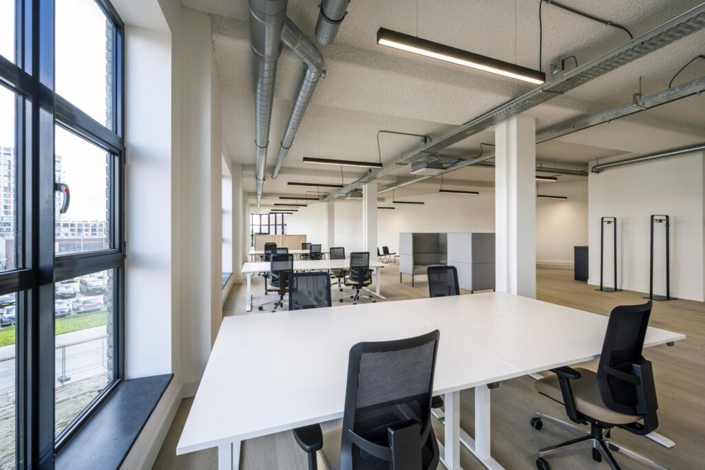 Modern open-plan office space with white desks, black chairs, and large windows providing natural light.