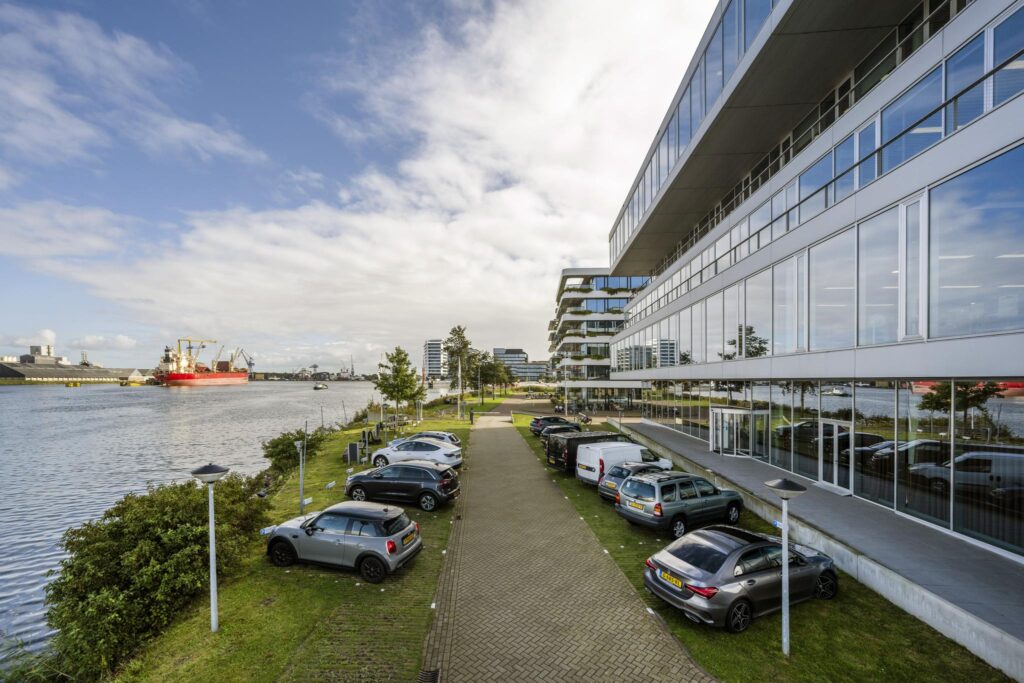 Modern office buildings along Moermanskade with parked cars and a view over the harbor featuring a red cargo ship.