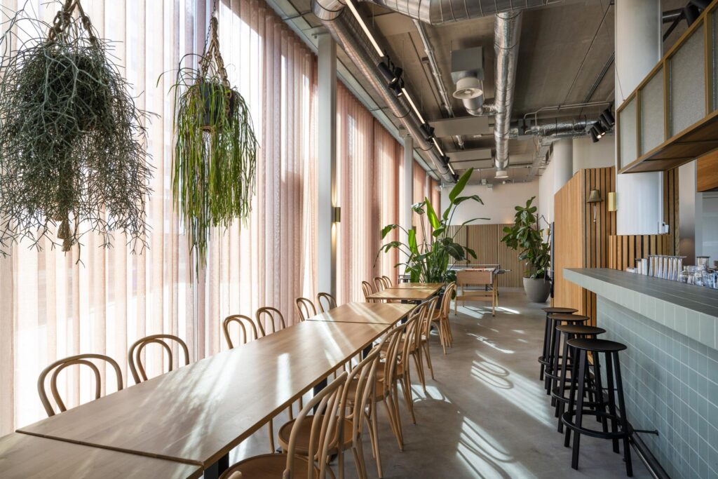 Modern communal workspace on Moermanskkade with long wooden tables, hanging plants, and a tiled bar.