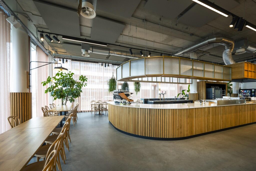 Modern cafeteria interior at Moermanskkade with a large wooden bar, dining tables, and natural light through sheer curtains.