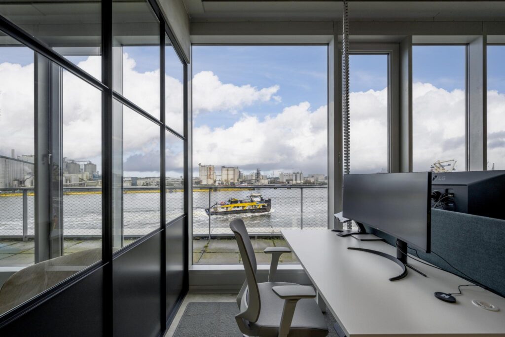 Modern office workspace with waterfront view overlooking an industrial area and a passing yellow ferry at Moermanskade.