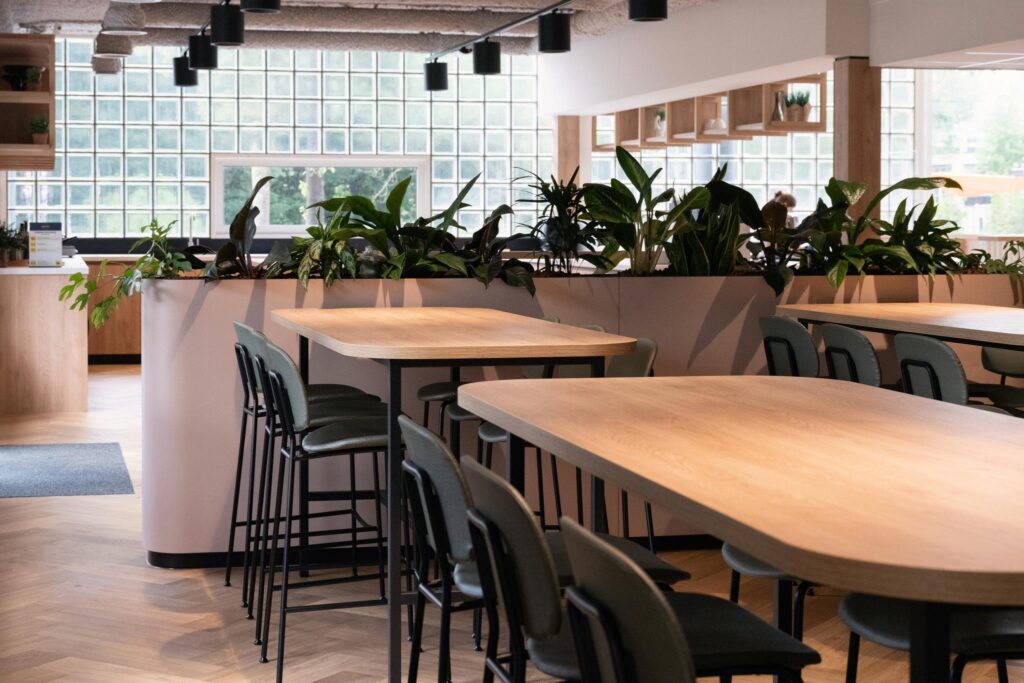 Modern communal workspace with wooden tables, green chairs, and lush indoor plants near glass block windows.
