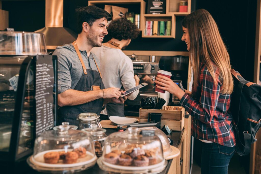 Barista smiling and taking an order from a customer holding a coffee cup at a café counter.