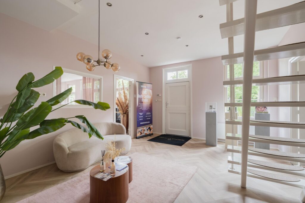 Modern waiting area of the EMS Body Contour Studio with soft pink walls, a curved beige sofa, spiral staircase, and promotional banner near the entrance.