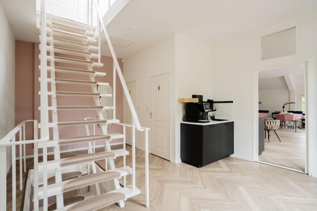 Modern hallway with a white staircase, coffee corner, and a view into a treatment room with pink-covered beds.