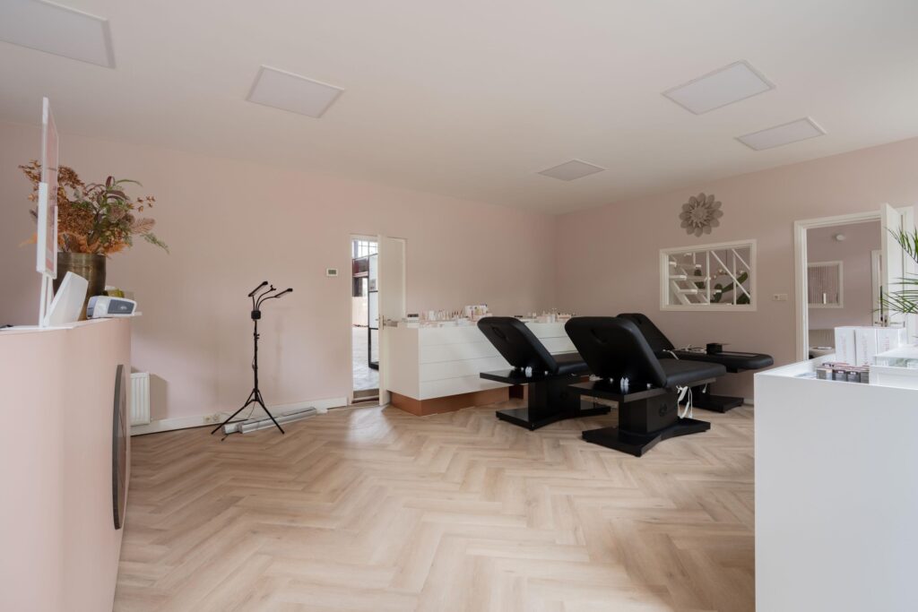Modern beauty treatment room with two black chairs, light pink walls, and a wooden herringbone floor.