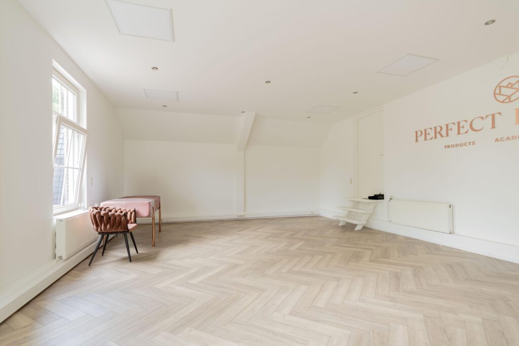 Bright, minimalistic room with herringbone wooden floor, a pink treatment table, and a modern chair near the window at Perfect Look Academy on Sloterweg.