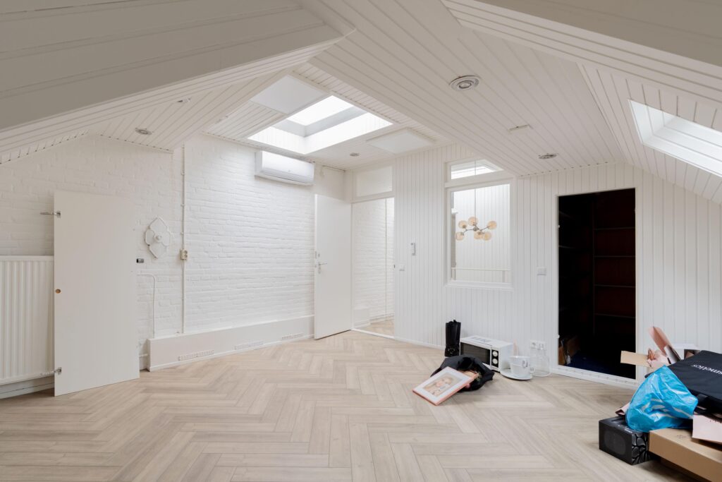 Bright attic room with white wooden walls, skylights, and scattered unpacked items on a light herringbone floor.