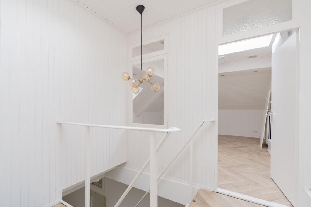 Bright white staircase landing with modern chandelier and doorway leading to attic room with herringbone floor.