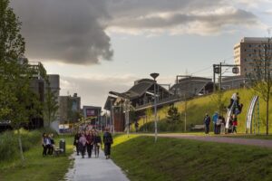 Groep mensen wandelt over een pad bij de Kuiperbergweg in Amsterdam-Zuidoost, met speeltoestellen en het station op de achtergrond.