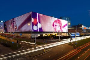 Groot LED-scherm op de gevel van de Johan Cruijff ArenA bij avond, gezien vanaf de Kuiperbergweg met lichtstrepen van passerend verkeer.