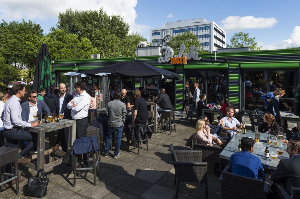 Mensen genieten van drankjes en gesprekken op het zonnige terras van restaurant De Zuiveringshal aan de Kuiperbergweg.