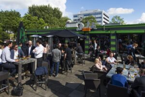 Mensen genieten van drankjes en gesprekken op het zonnige terras van restaurant De Zuiveringshal aan de Kuiperbergweg.