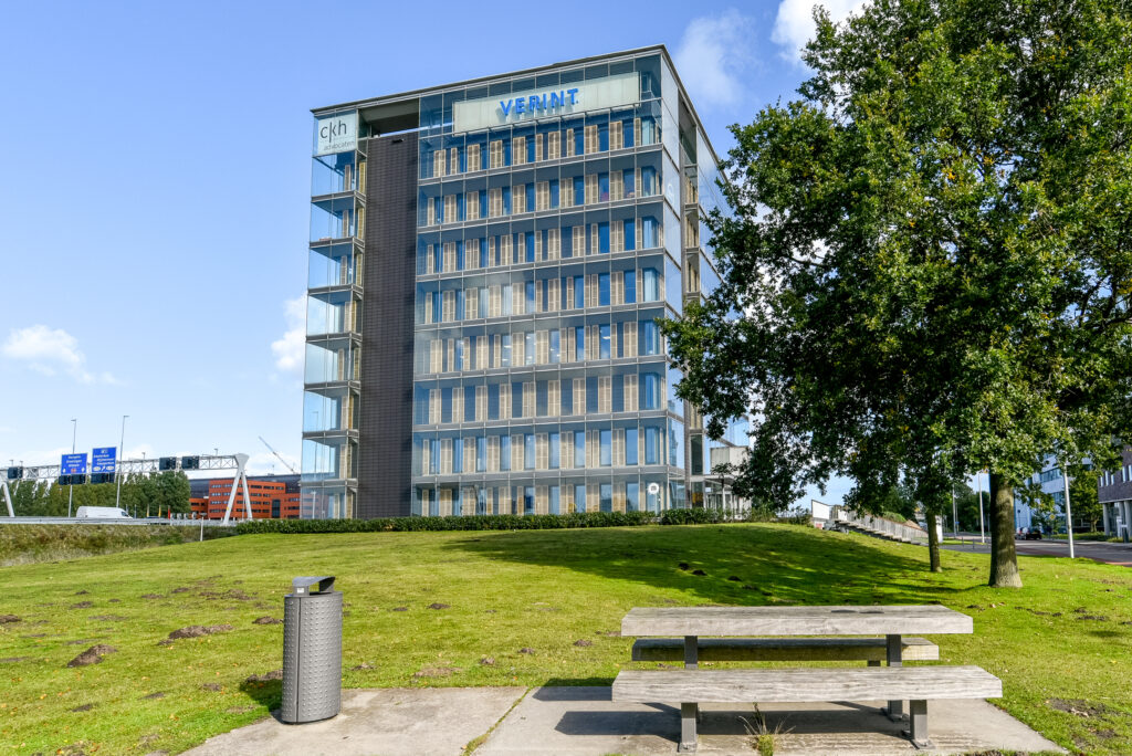 Modern office building on Paasheuvelweg with Verint and CKH Advocaten signage, viewed from a grassy area with a bench and tree.