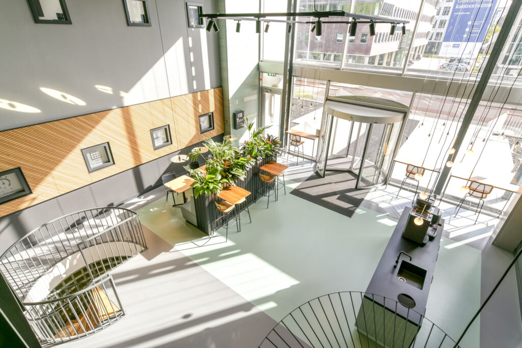 Bright and modern lobby interior at Paasheuvelweg with seating areas, indoor plants, spiral staircase, and large windows.