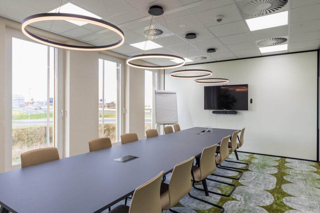 Modern conference room on Paasheuvelweg with a large table, beige chairs, circular ceiling lights, and a wall-mounted screen.