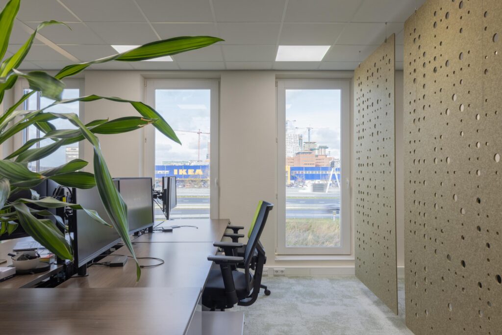 Modern office workspace with desks and monitors, featuring a view of the IKEA store on Paasheuvelweg through large windows.
