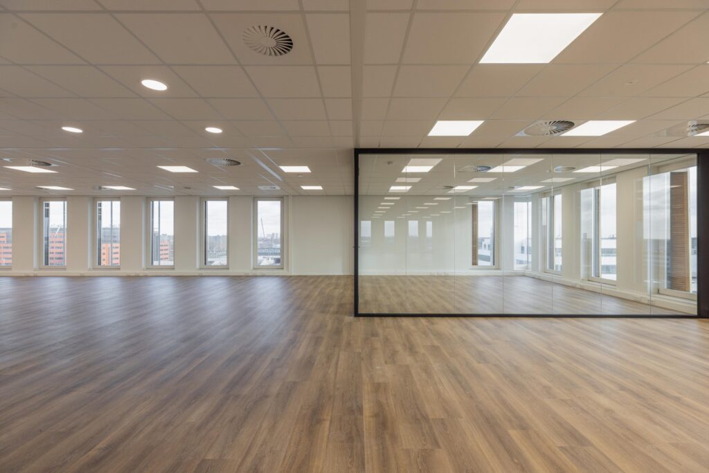 Modern empty office space at Paasheuvelweg with large windows, glass walls, and wooden flooring.