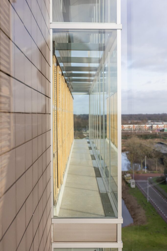 Glass corridor with wooden blinds on the Paasheuvelweg building, overlooking a road and waterway.