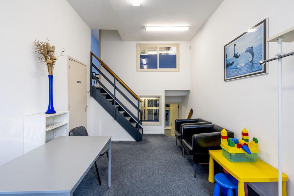 Waiting area with black chairs, a yellow children's play table with building blocks, a staircase, and a dolphin-themed wall poster.