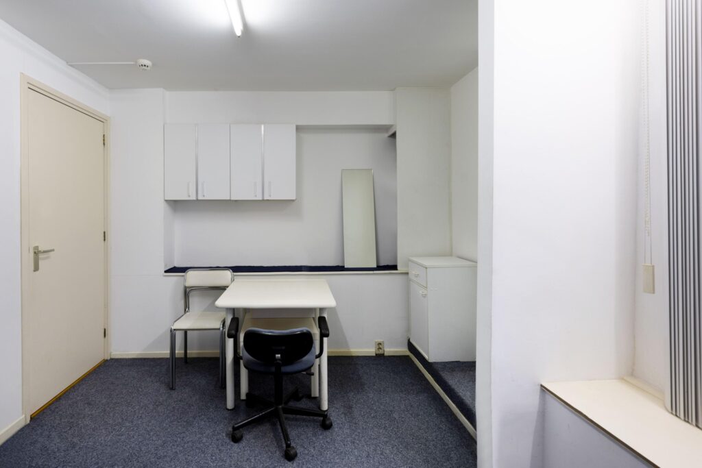 Small, minimalistic office space with two chairs, a white table, wall cabinets, and a mirror against the back wall.