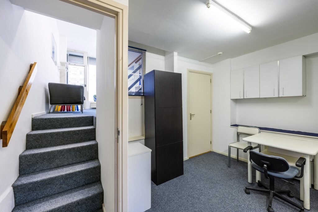 Small office space with carpeted floor, white walls, a desk with chairs, cabinets, and a staircase leading to a seating area.