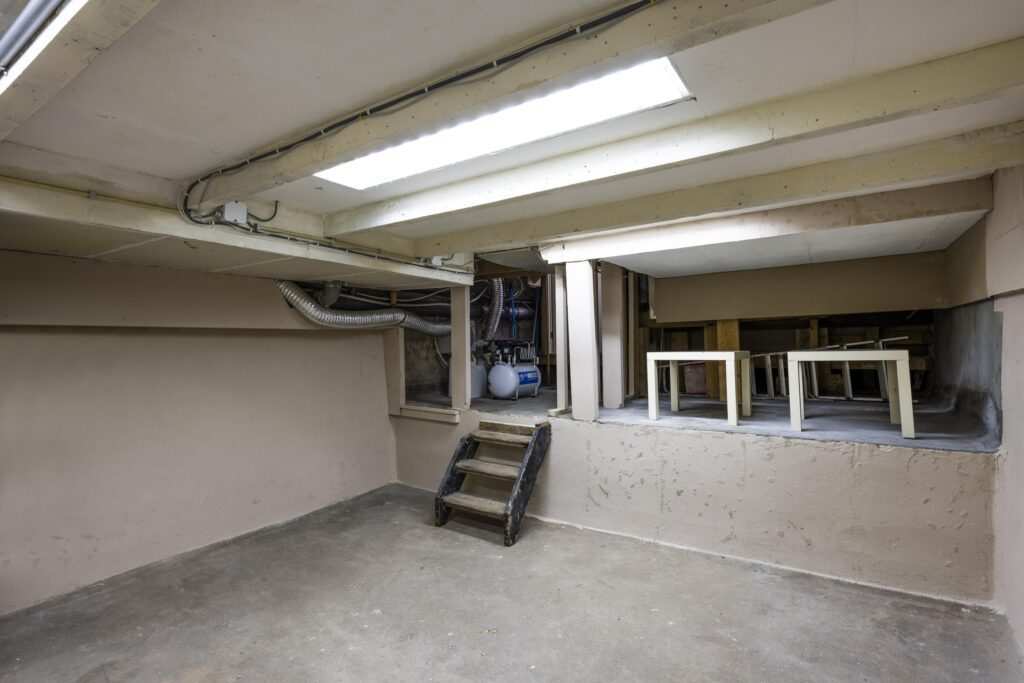 Basement space with concrete floor, exposed piping, and a small staircase leading to a raised storage area with tables.