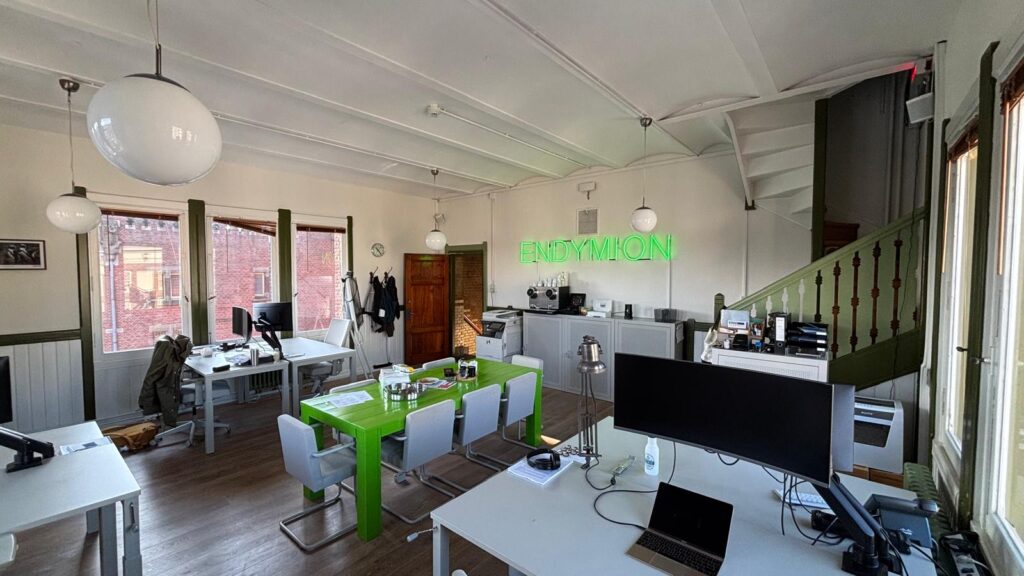 Modern office interior with white desks, a bright green meeting table, and a neon "ENDYMION" sign on the wall.