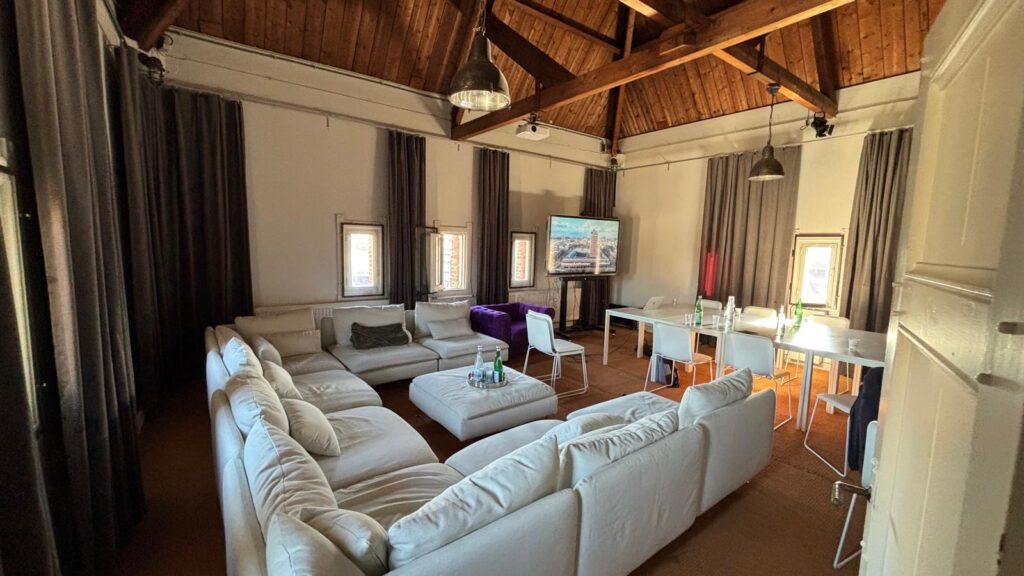 Spacious meeting room on Damrak with a large white sectional sofa, dining table, TV screen, and high wooden ceiling.