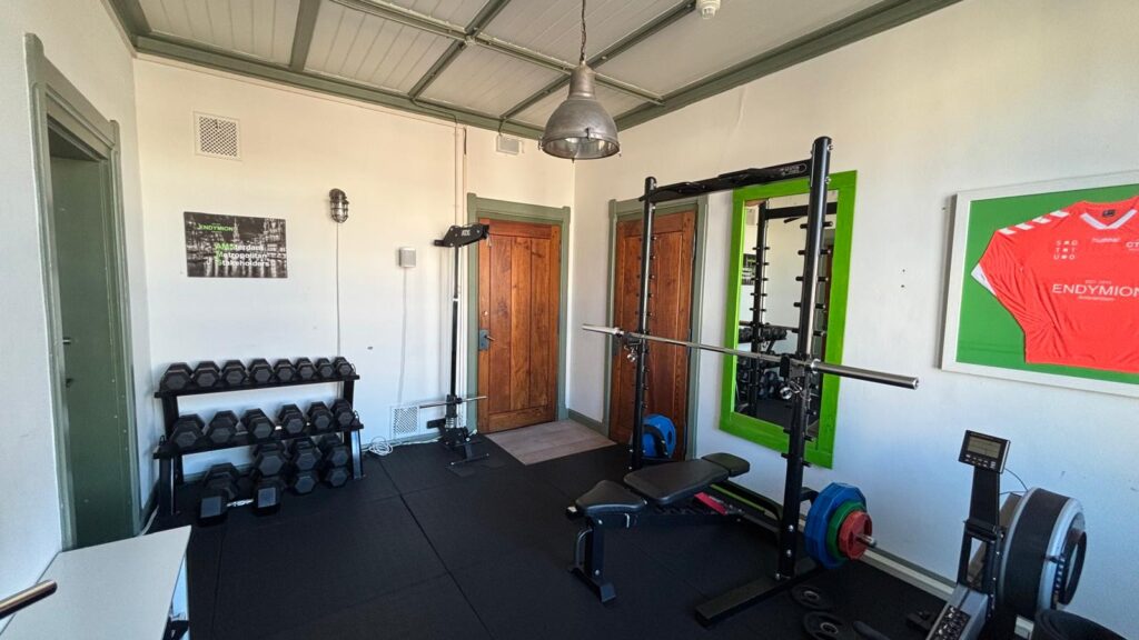 Small home gym with weightlifting equipment, dumbbells, and a framed red Endymion Amsterdam sports jersey on the wall.