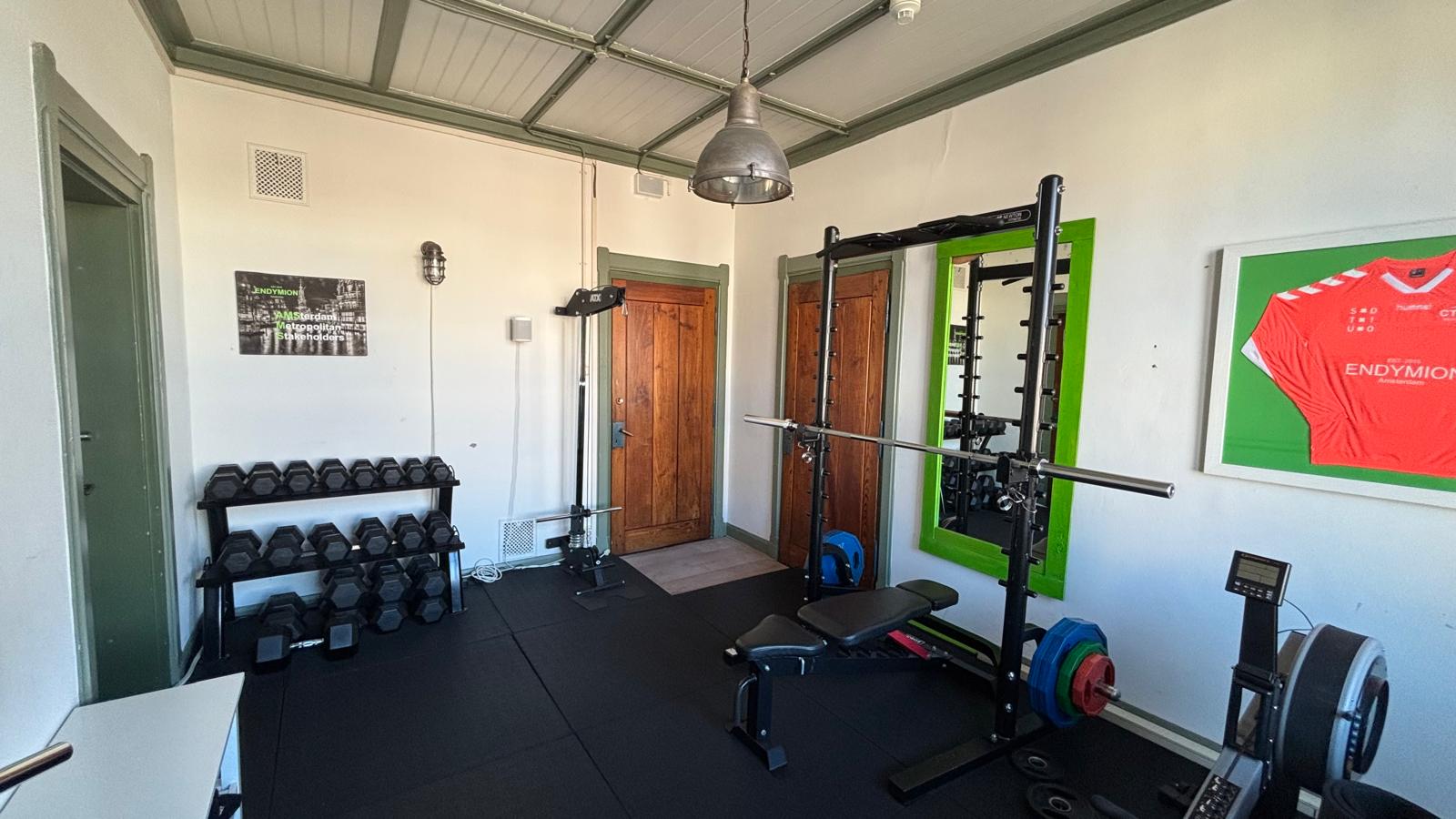 Kleine fitnessruimte met krachttrainingsapparatuur, gewichten en een roeimachine bij Damrak 246 in Amsterdam.