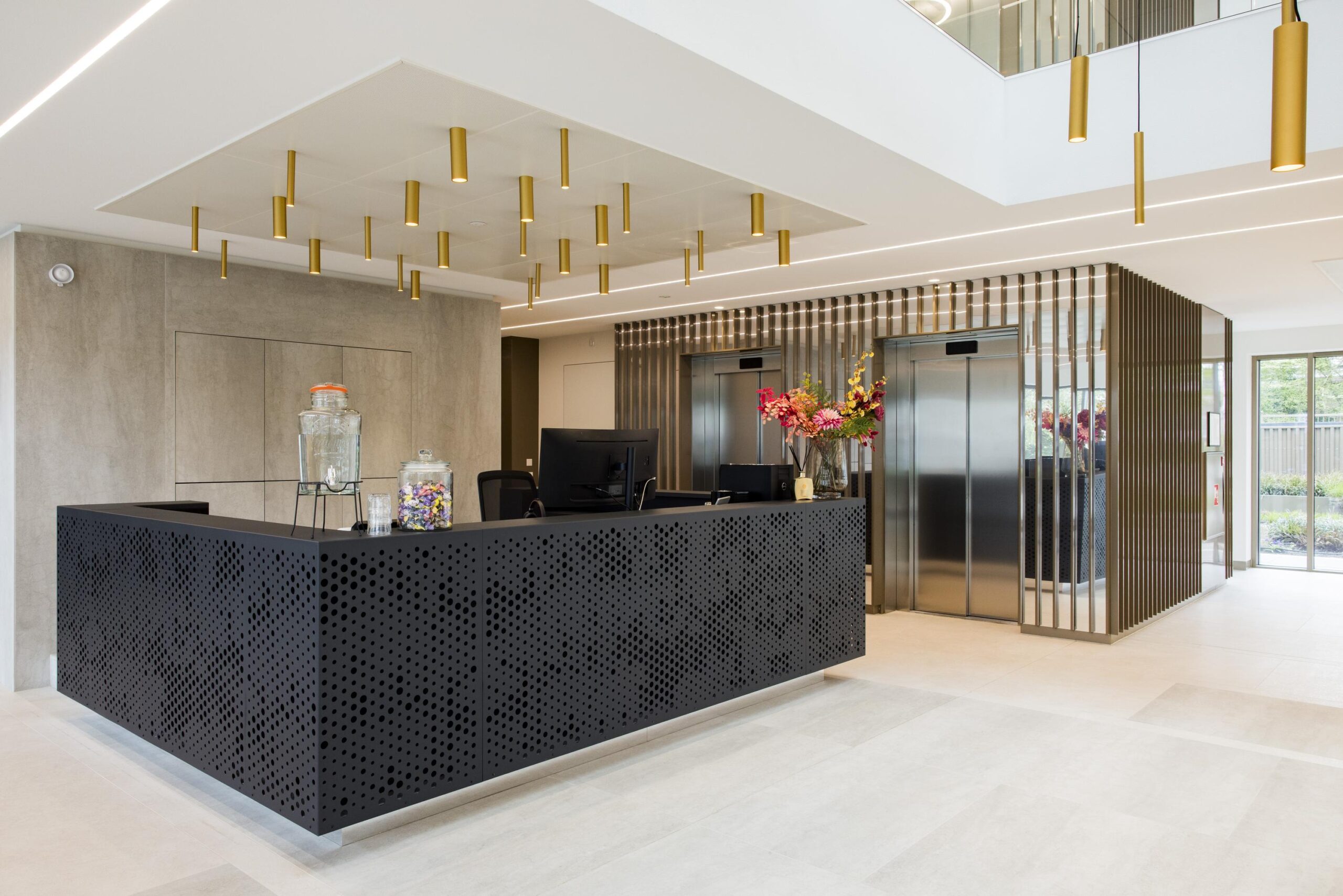 Modern office lobby with a black perforated reception desk, decorative lighting, and stainless steel elevators.