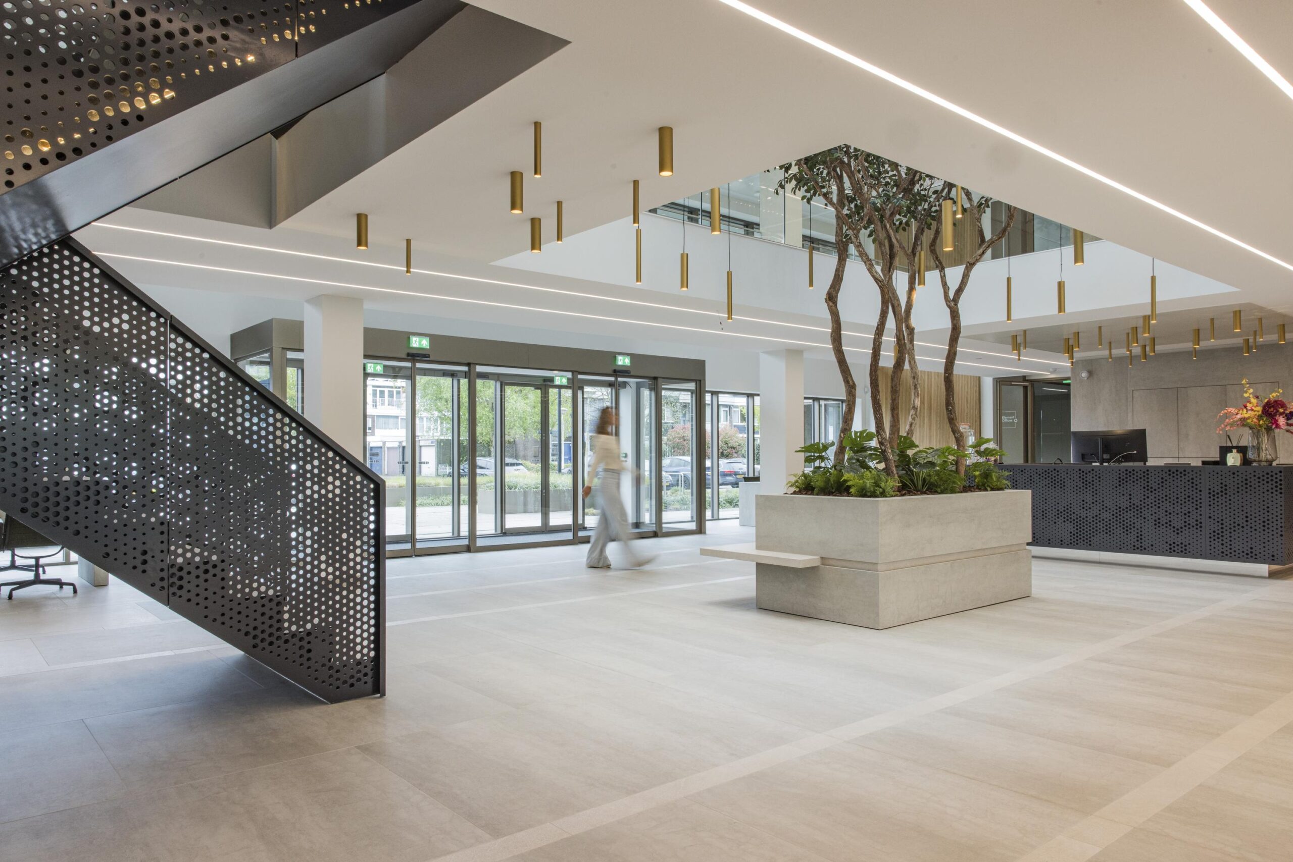 Modern office lobby with a perforated metal staircase, indoor trees, and a reception desk.