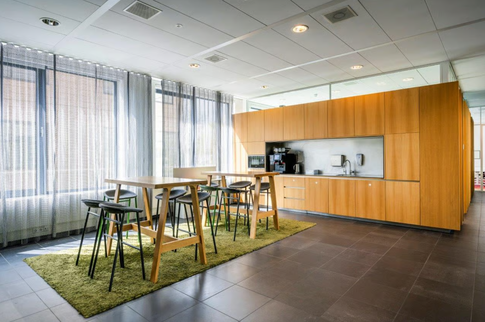 Modern office kitchen with wooden cabinets, high tables, and bar stools on a green rug.