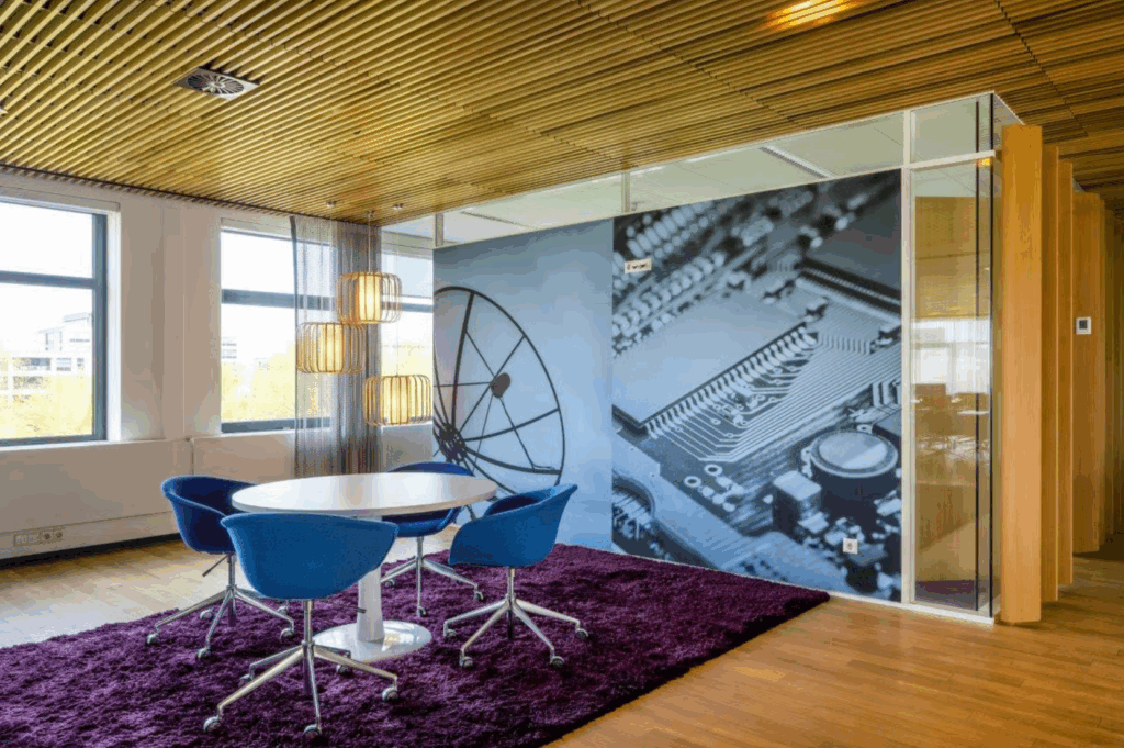 Modern office seating area with blue chairs, a round table, and a wall mural featuring a satellite dish and microchip design.