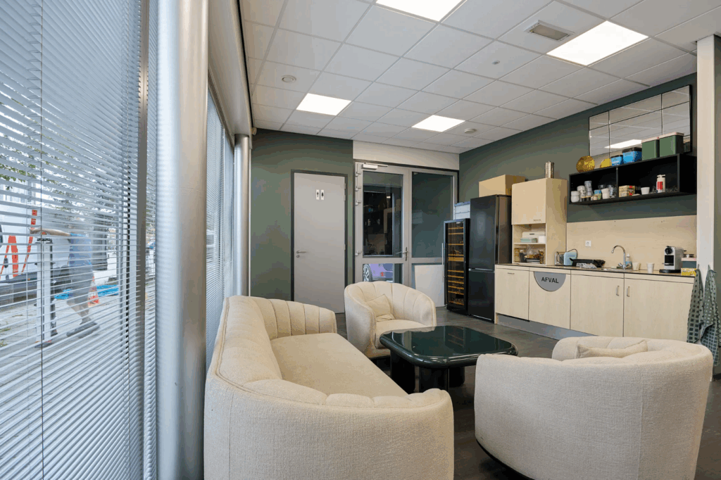 Modern staff kitchen and lounge area with beige seating, coffee station, and large windows with blinds.