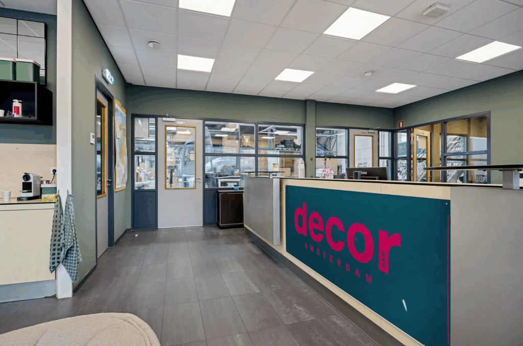 Modern reception area of Decor Amsterdam with a teal and pink front desk and view into the warehouse through glass windows.
