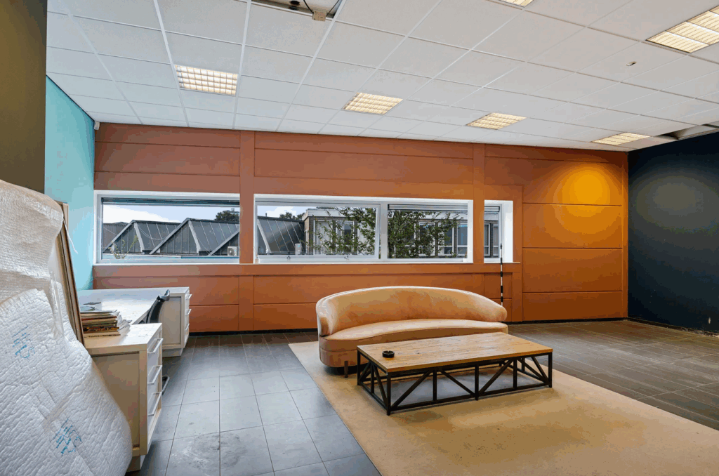 Modern office lounge area with a light pink curved sofa, wooden coffee table, and large windows overlooking rooftops.