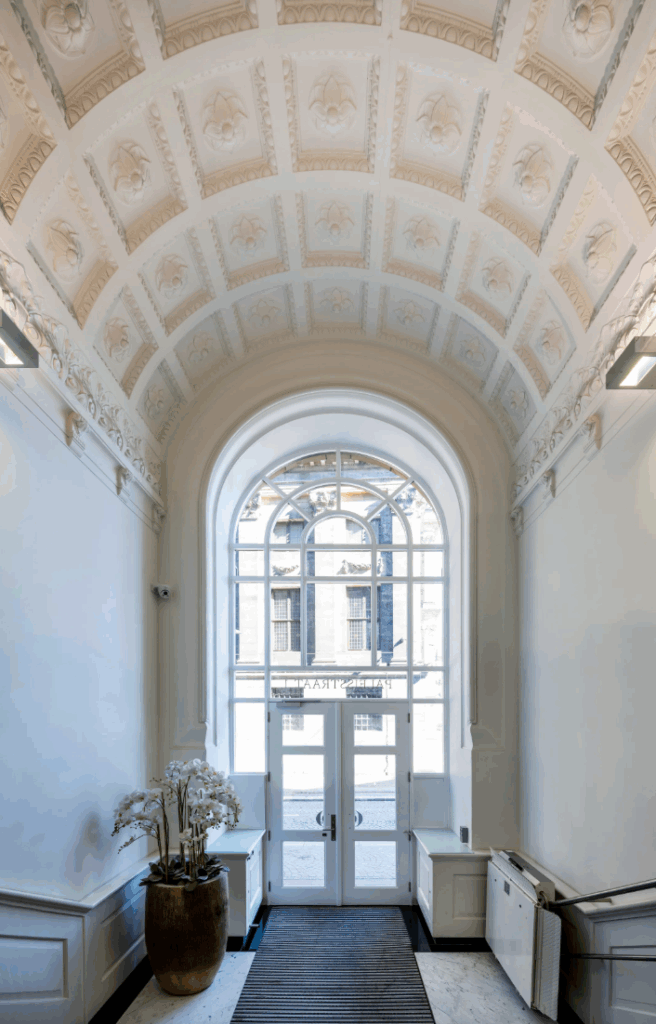 Elegante gewelfde entreehal met sierlijke plafonddetails en uitzicht op de Paleisstraat door grote glazen deuren.