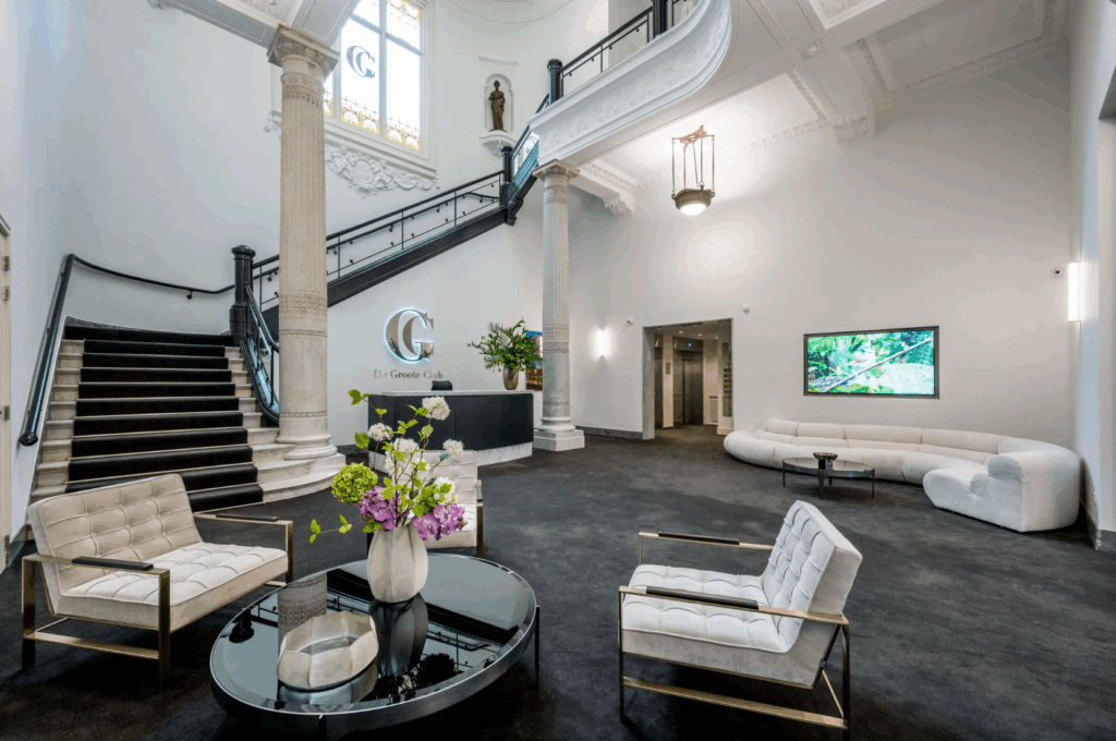 Elegante entreehal van De Groote Club met moderne zitplaatsen, een grote trap en decoratieve zuilen.